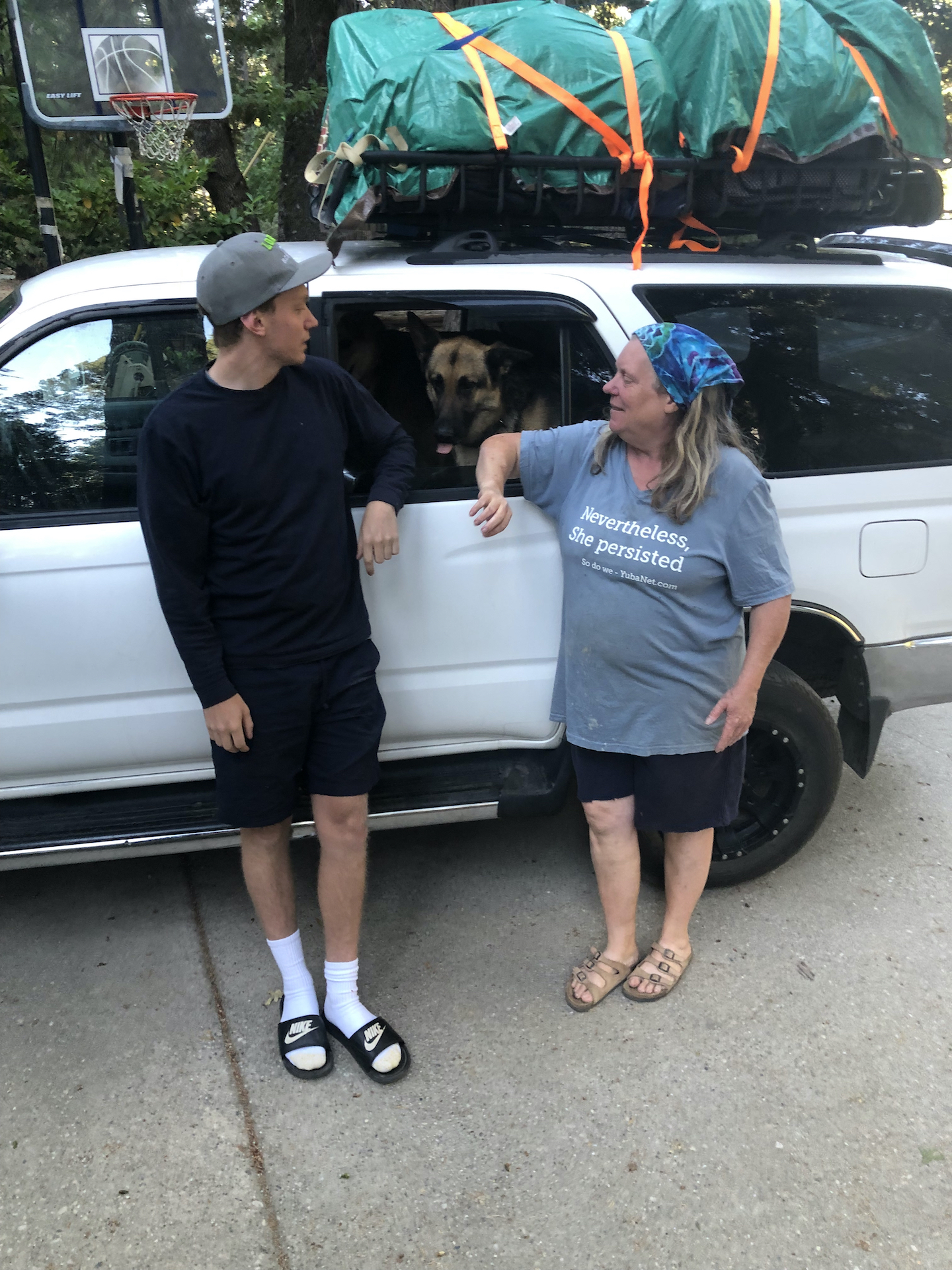 A woman and her son gaze at their German Shepherd who is poking his head out the door of an SUV they are leaning against.