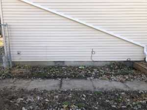 An exterior wall of a home with a diagonal drain spout. Below, a patch of dirt separated by a sidewalk.