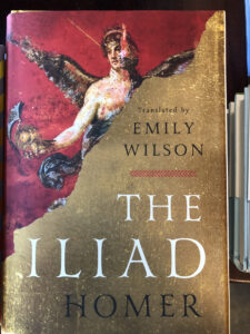 A hard cover book, The Iliad, translated by Emily Wilson. The books is mostly tan with some red upper left and a painting of an ancient Greek. 