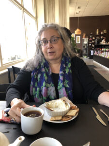 A lovely gray haired woman in black with a blue floral scarf holds a cup of tea. Beneath her on the table is a bowl of goat cheese sitting on a plate with several crackers.