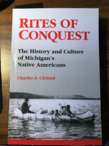 A gray book with red letters that say Rites of Conquest and black letters beneath that say The History and Culture of Michigan's Native Americans. Below that the author's name in red, Charles E Cleland.