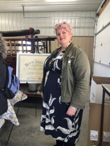 A tall, thick, blond woman in a blue and white print dress and green army jacket stand in front of an informational sign and smiles at the camera.