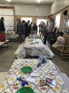 A crowd of people enters the doorway in background. Large tables with plates, spoons, and seed packets in foreground.