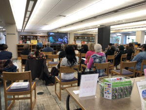 A small group of people sit-in chairs facing a large screen viewing a film. They are sitting in a library.