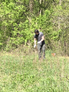 A tall white man in gray clothing pushes a piece of rebar into the soil. He is standing in meadow.