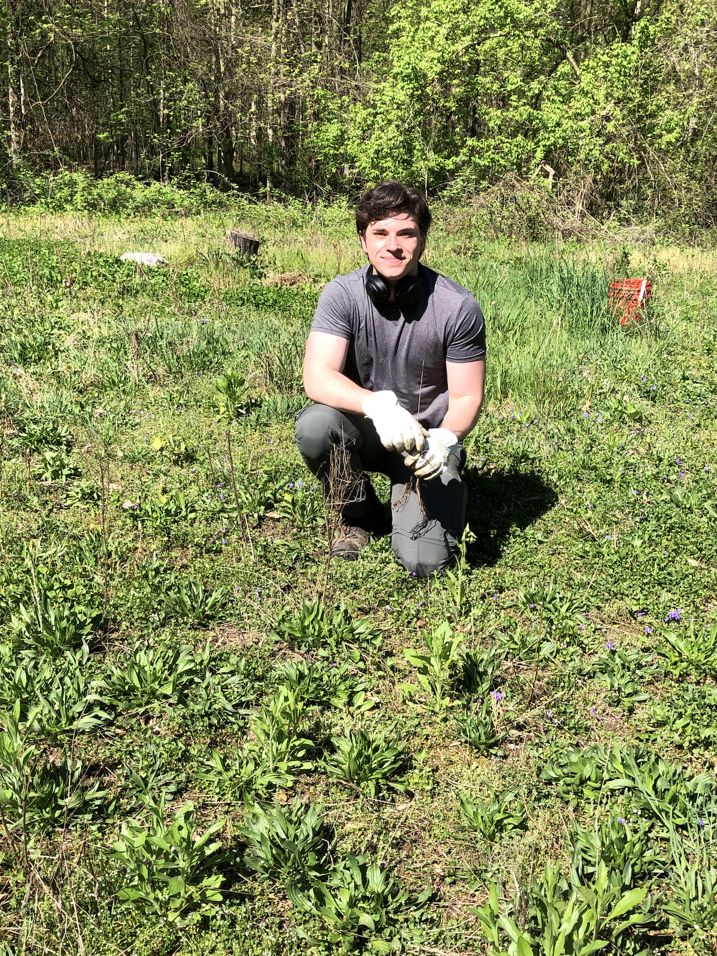 A young white man squats in a meadow. He's dressed in gray with white gloves. He is smiling.