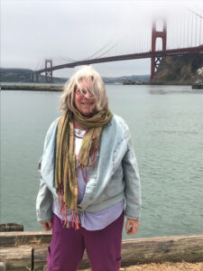 A gray haired woman stand along the San Francisco Bay. Her hair is wind-tossed. The Golden Gate Bridge is Behind her.