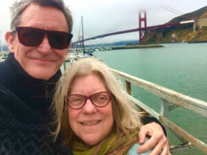 A tall man in sunglasses has his arm around a shorter woman in regular glasses. The Golden Gate Bridge is in the background.