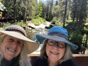 Two women, wearing sun has, stand on a wooden bridge overlooking a river with granite boulders and conifers beside it. The women are smiling.